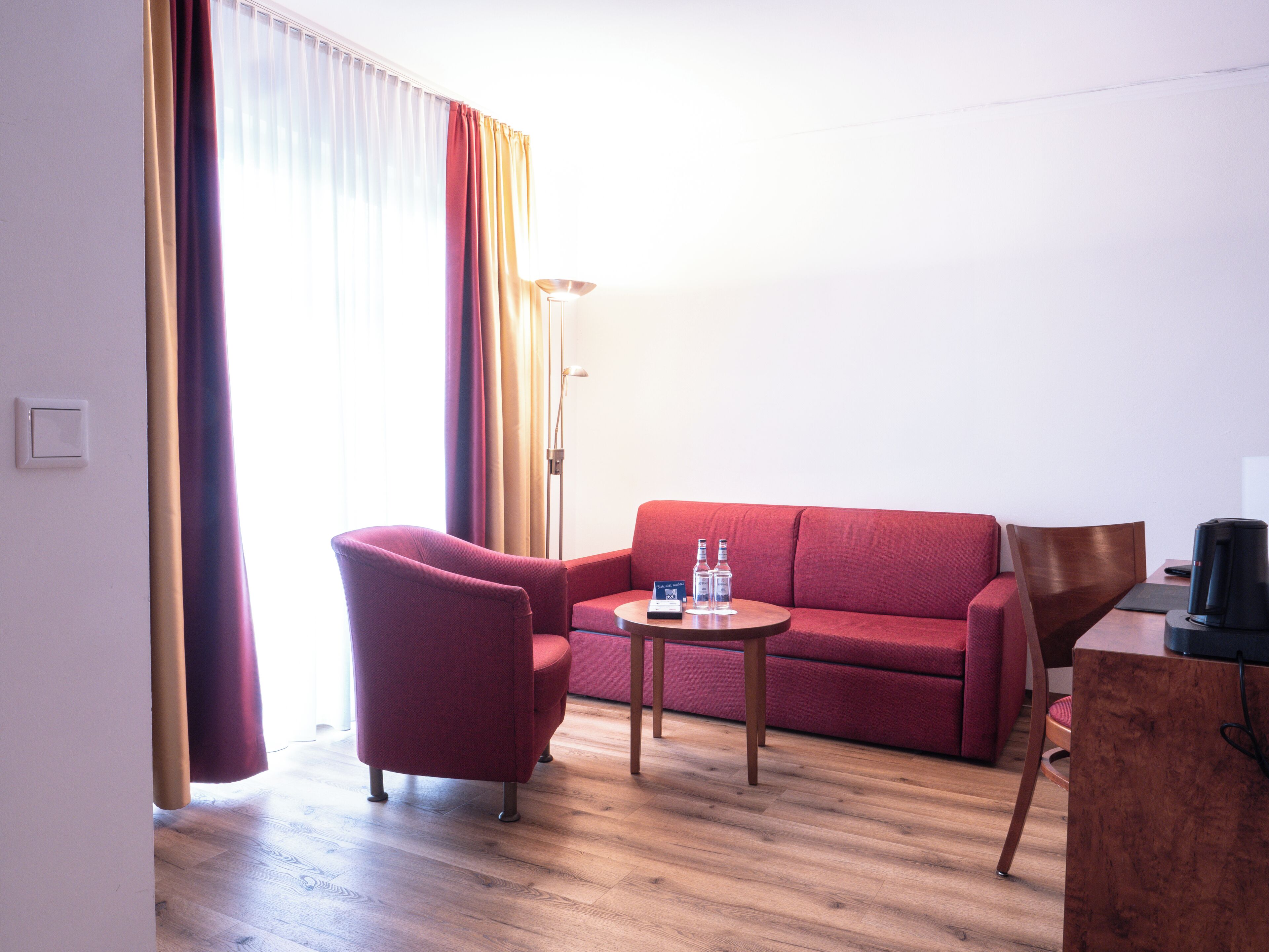 Ett hotellrum med en röd soffa, ett träbord, en röd fåtölj, ett skrivbord med en svart kaffebryggare och en golvlampa.