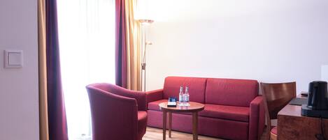 A hotel room with a red sofa, a wooden coffee table, a red armchair, a desk with a black coffee maker, and a floor lamp.