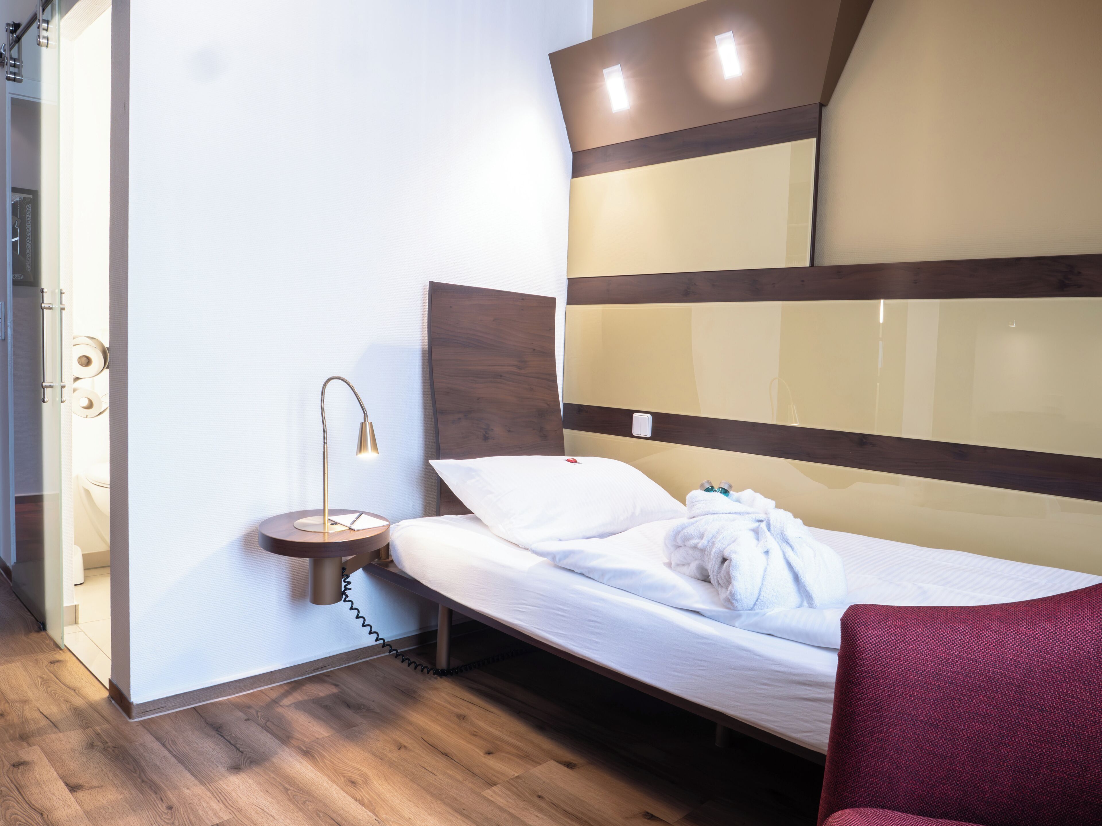 A modern hotel room with a wooden floor, a bed with white bedding, a bedside table with a lamp, and a red chair.
