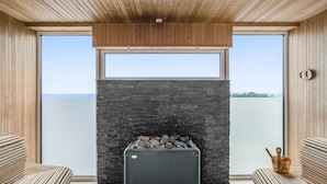 Un intérieur moderne avec une cheminée en pierre, un plafond en bois et de grandes fenêtres donnant sur un plan d’eau.