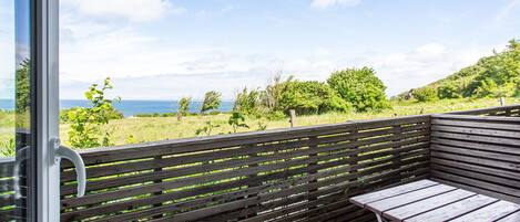 Un balcón con banca y mesa de madera, con vista a un área verde con árboles y al mar.