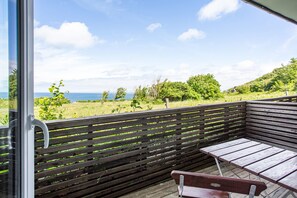 Een balkon met een houten bankje en tafel, met uitzicht op een grasveld met bomen en de zee.