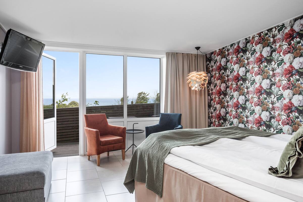 Ein Schlafzimmer mit Bett, Fernseher, Sofa, zwei Sesseln, einem kleinen Tisch und einem Balkon mit Aussicht.