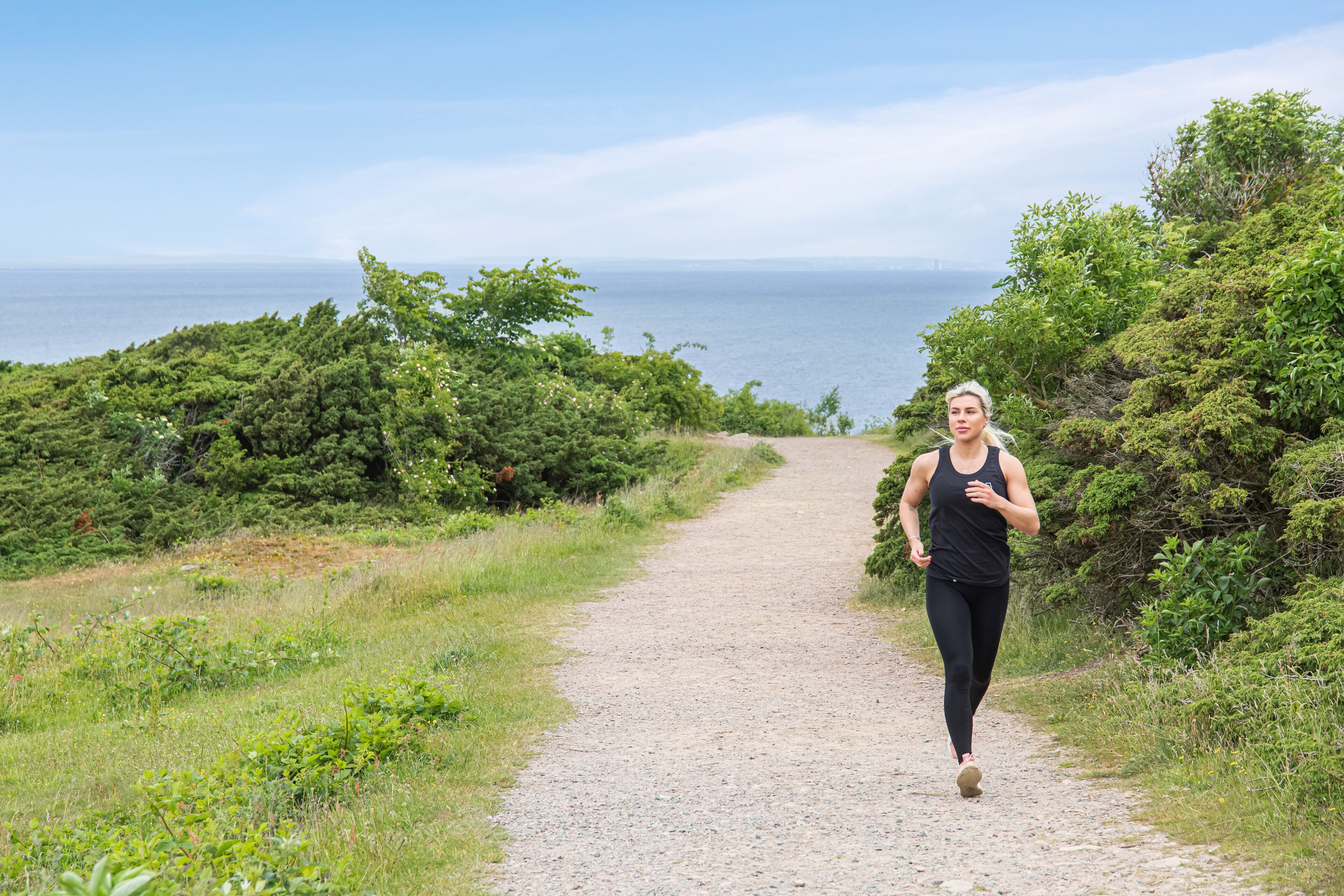 A person jogging on a gravel path surrounded by greenery and overlooking a body of water.