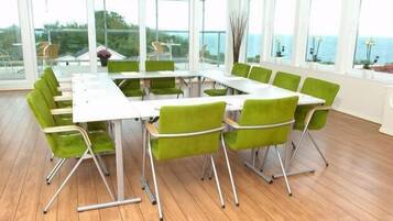 A conference room with a long table and green chairs, large windows with a view of the sea, and a wooden floor.
