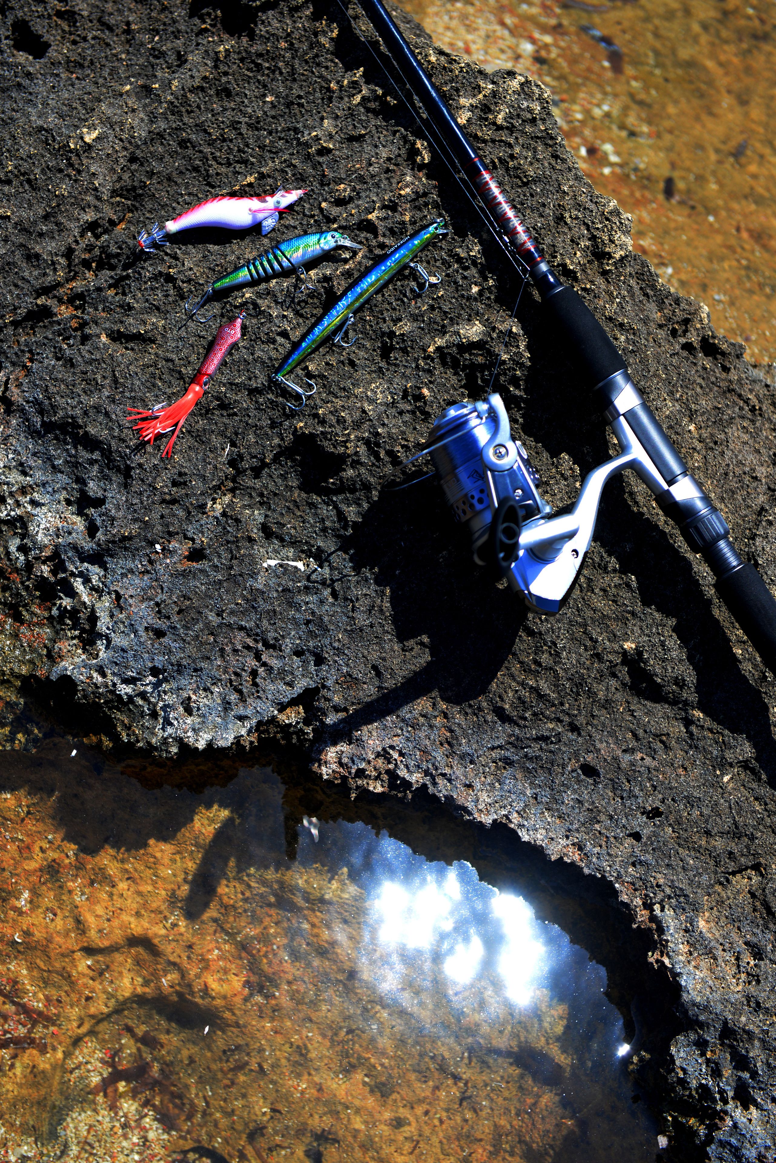A fishing rod, several lures, and a fishing reel on a rock by a body of water.