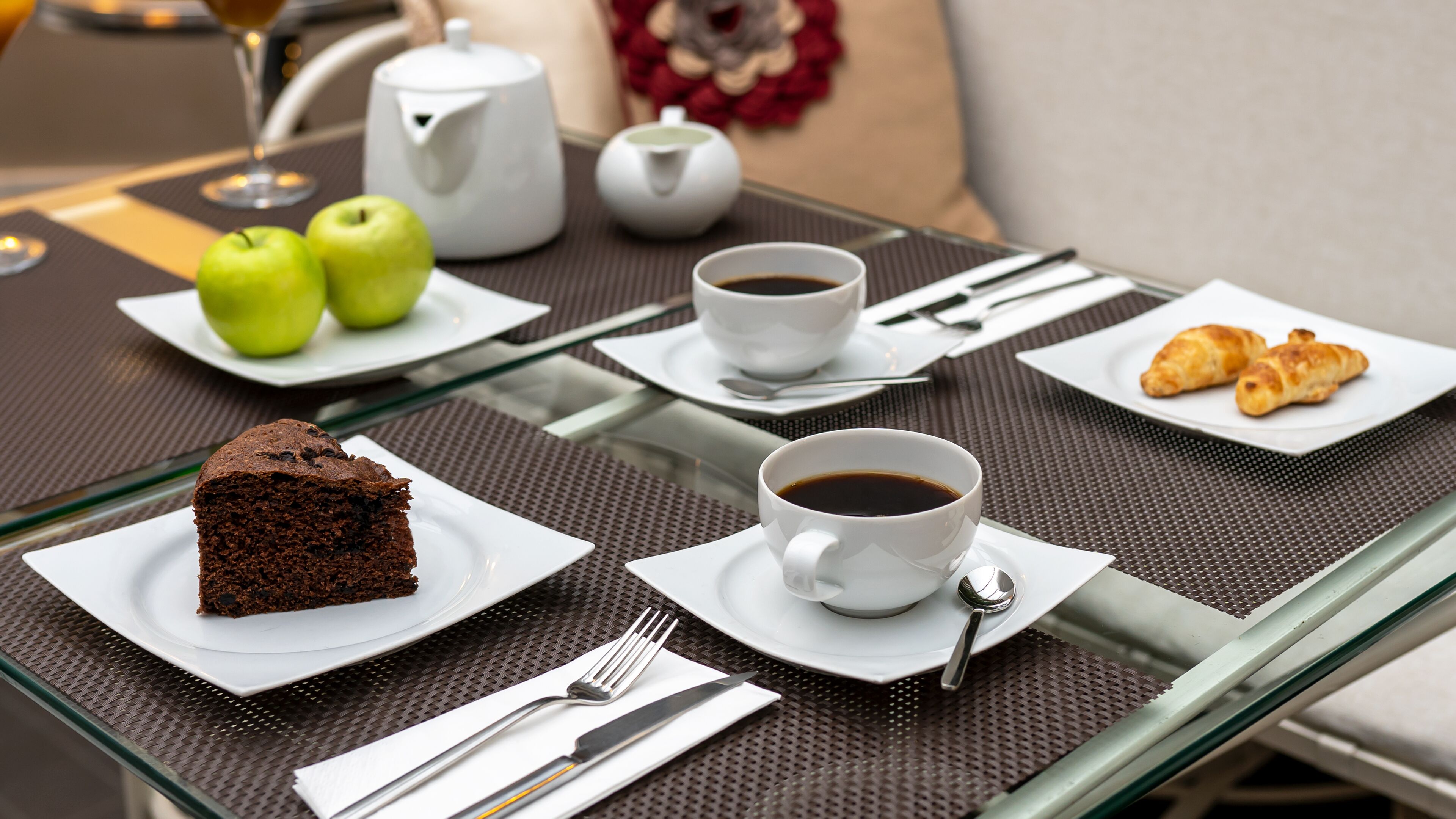 En frukostuppdukning med en bit chokladkaka, två koppar kaffe, gröna äpplen, croissanter och en tekanna på ett glasbord.