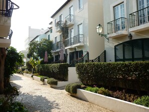 Calle adoquinada bordeada por setos cuidados y una hilera de edificios blancos con balcones.