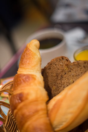 A basket containing a croissant and a slice of bread, with a cup of coffee and a small container of butter in the background. - Palma Hotel (Paris)