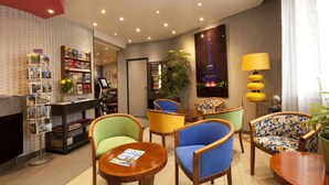 Un vestíbulo de hotel con una mesa de recepción, una estantería con folletos turísticos, sillas coloridas, una planta grande en maceta y una lámpara de pared.