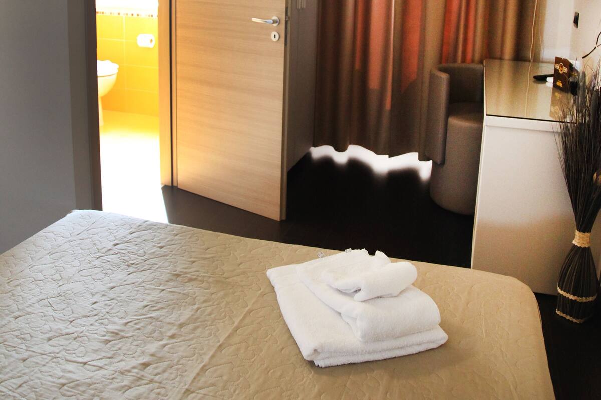 Habitación de hotel con cama, escritorio, silla y baño visible a través de una puerta abierta.