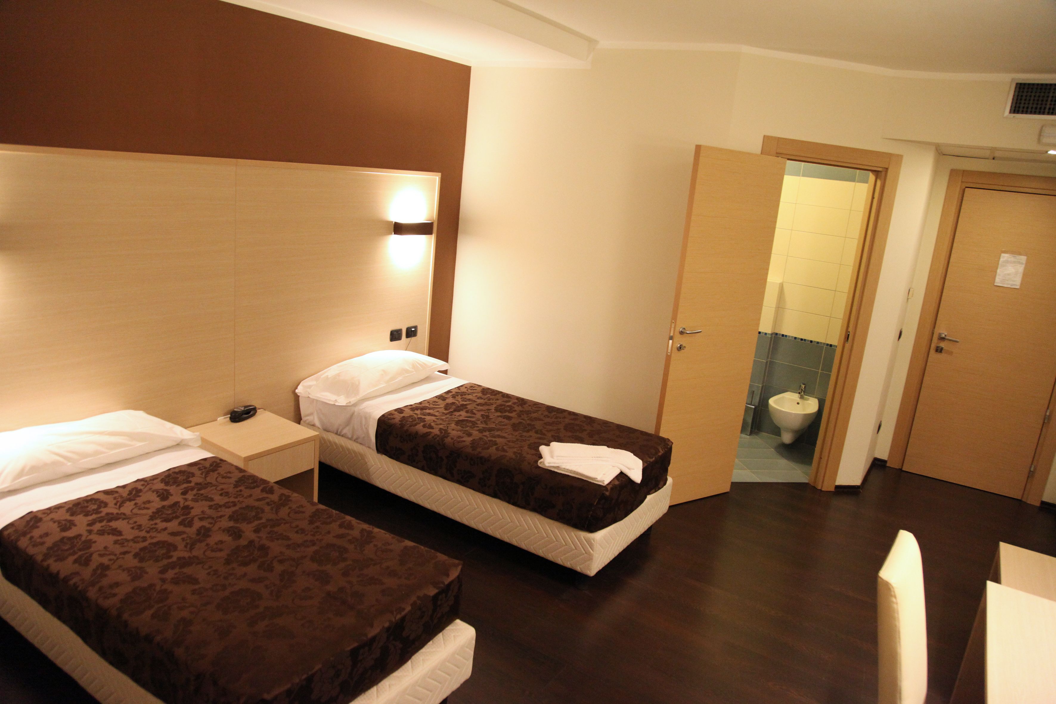 A hotel room with two beds, a bedside table with a phone, a wall-mounted light, and a door leading to a bathroom.