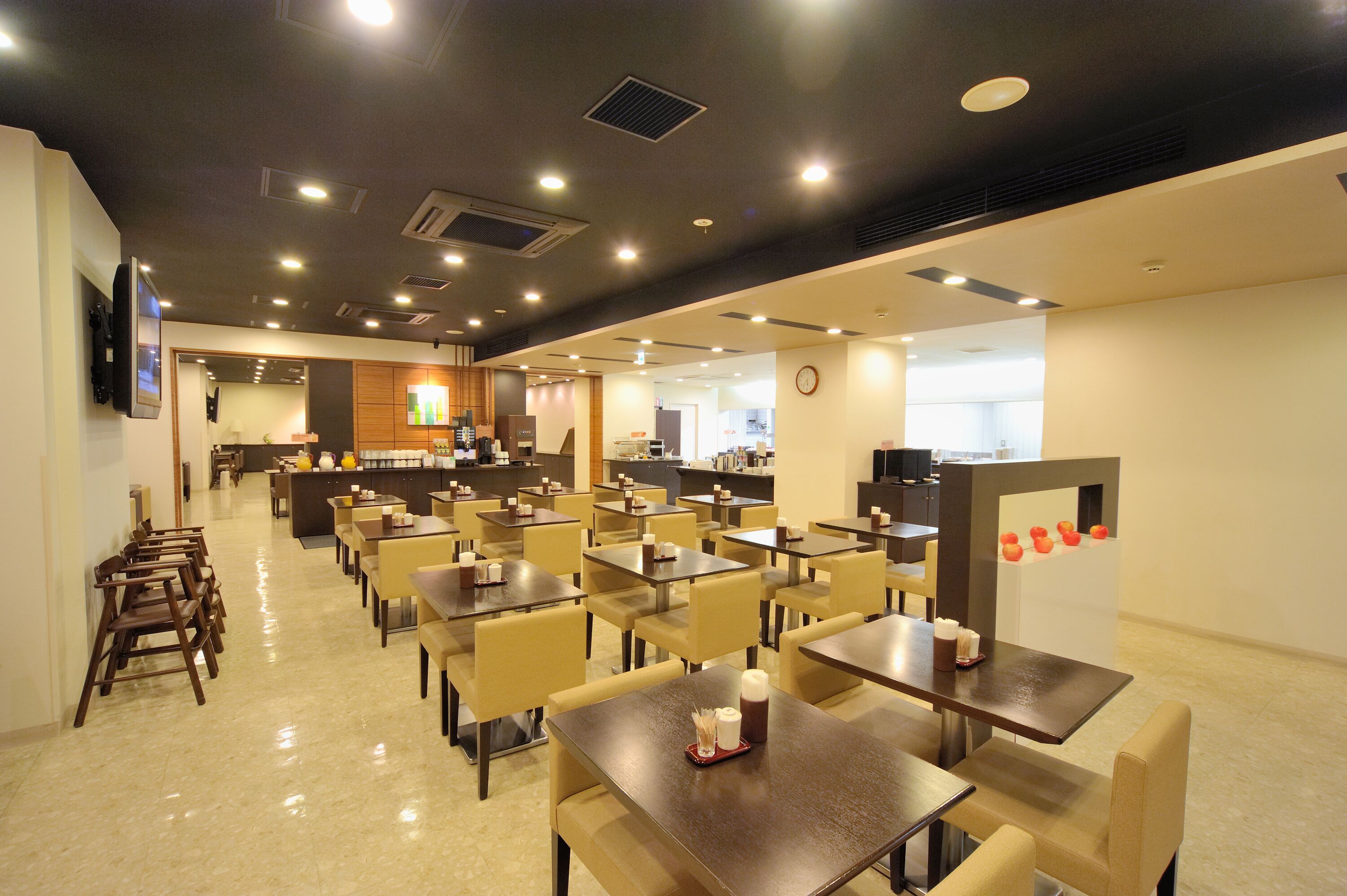 A spacious dining area with multiple tables and chairs, a kitchen in the background, and a clock on the wall.