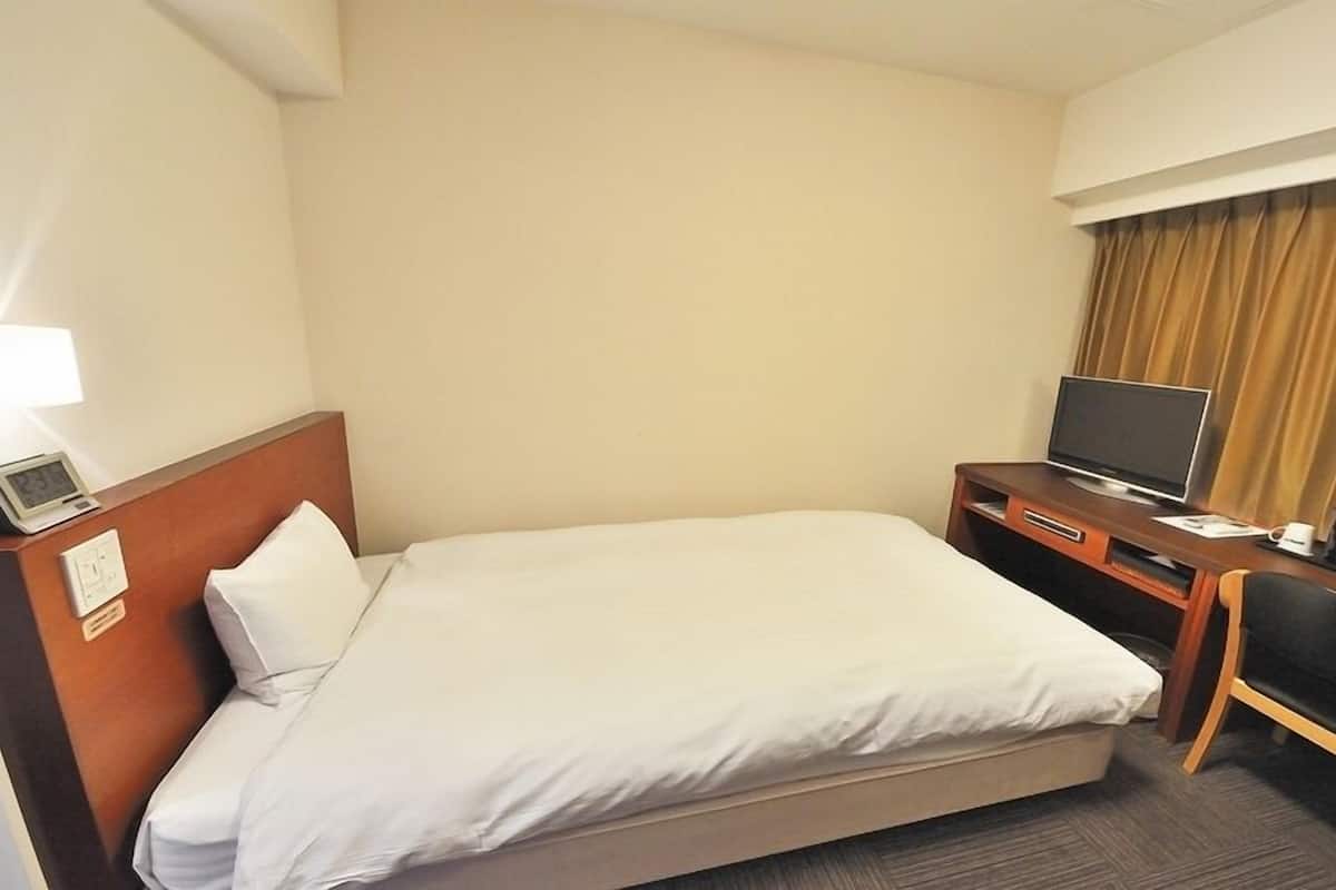 A hotel room with a bed, a desk with a TV, a chair, a clock, and a window with curtains.