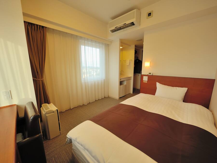 A hotel room with a large bed, a desk, a chair, and a trash bin.