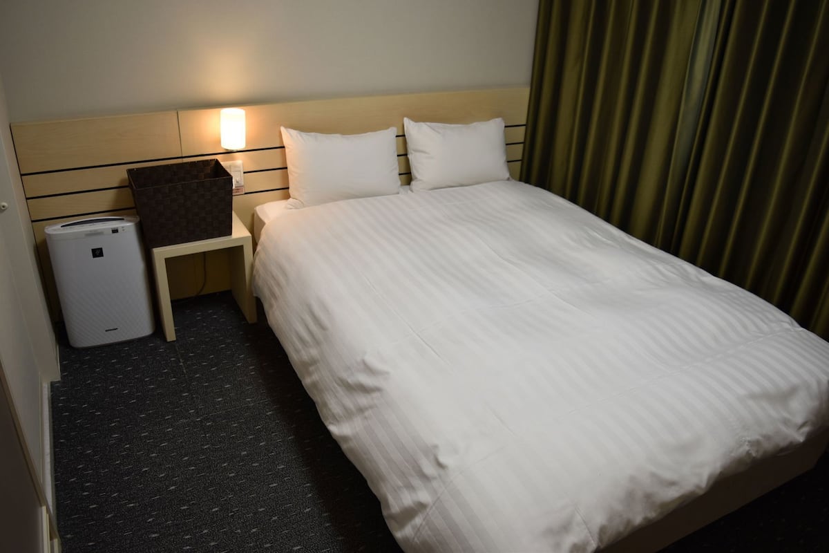 Ett hotellrum med en säng, ett nattduksbord, en lampa, en papperskorg och en väggmonterad värmare.