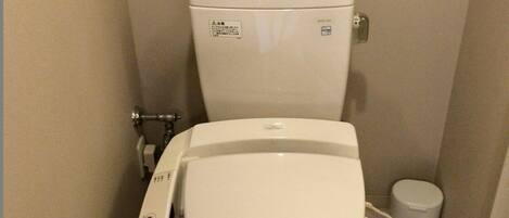 A compact bathroom with a white toilet, a bidet attachment, and a small trash bin.