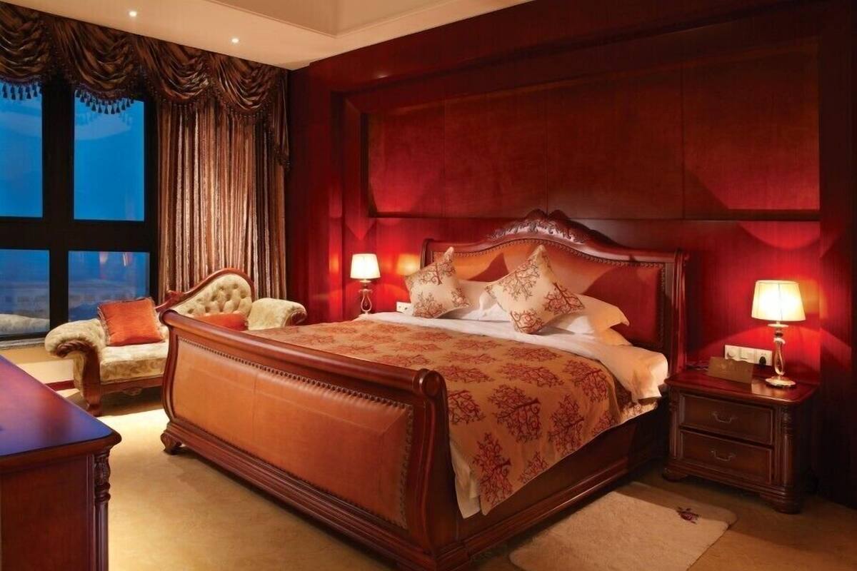 A large, ornate bed with a wooden frame and a red headboard, flanked by two nightstands with lamps, in a room with a large window and a view of the outdoors.