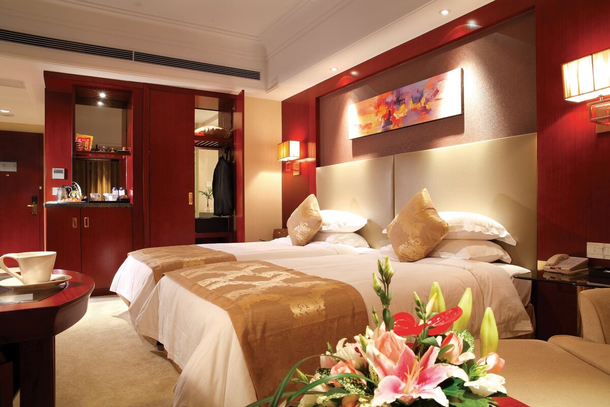 A hotel room with two beds, a sitting area with a table and chairs, a minibar, and a large painting on the wall.