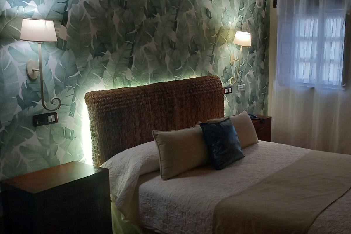 Un llit ben fet amb capçalera, dues làmpades de nit i una finestra amb cortines translúcides.