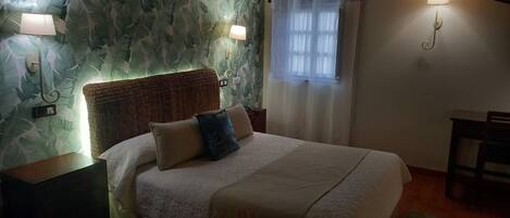 Cama bem arrumada com cabeceira, duas luminárias de apoio e janela com cortinas leves.