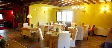 A dining area with tables set for four, wooden beams on the ceiling, and a large window with curtains.