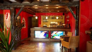 A bar area with a red wall, wooden ceiling beams, and a tiled floor. There are wicker chairs and a bar counter with bottles and glasses. - La Casona de Revolgo (Santillana del Mar)