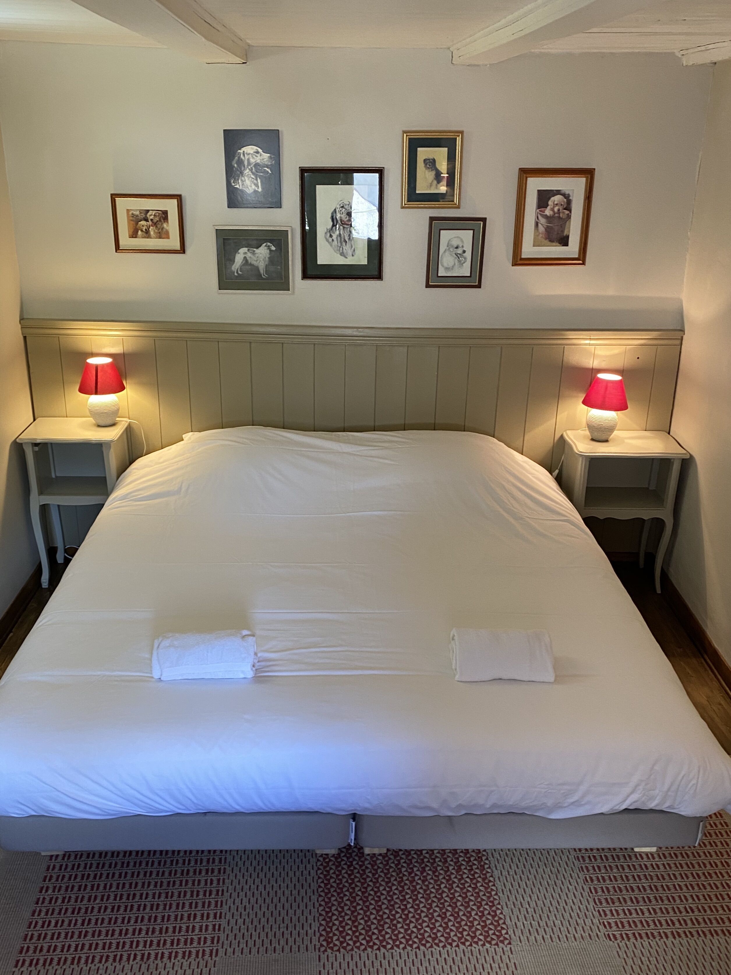 A neatly made bed with white linens and two towels, flanked by two bedside tables with lamps, in a room with a wooden headboard and framed pictures on the wall.