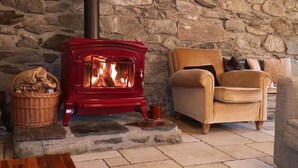 A cozy room with a stone wall, a red wood-burning stove, a wicker basket, and a beige armchair. - Maison Caramel (Landry)