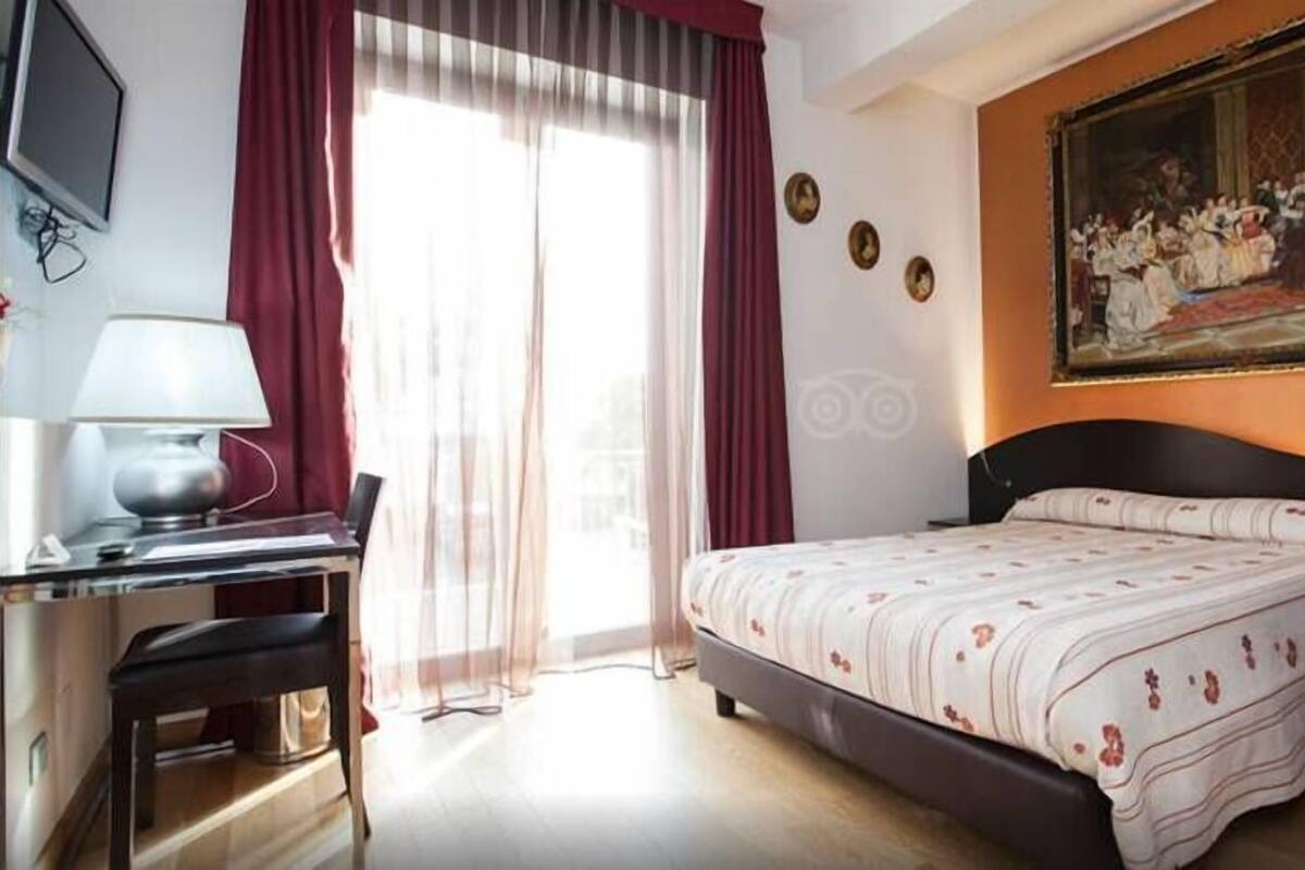 A hotel room with a bed, a desk, a chair, a TV, a painting on the wall, and a window with curtains.