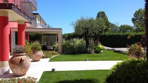 A modern building with a red facade, a covered patio area, and a well-maintained garden with various plants and a lawn. - La Grotta Hotel (Verona)