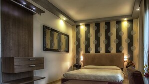 A modern bedroom with a large bed, a TV cabinet, a patterned wall, and a nightstand with a lamp. - La Grotta Hotel (Verona)