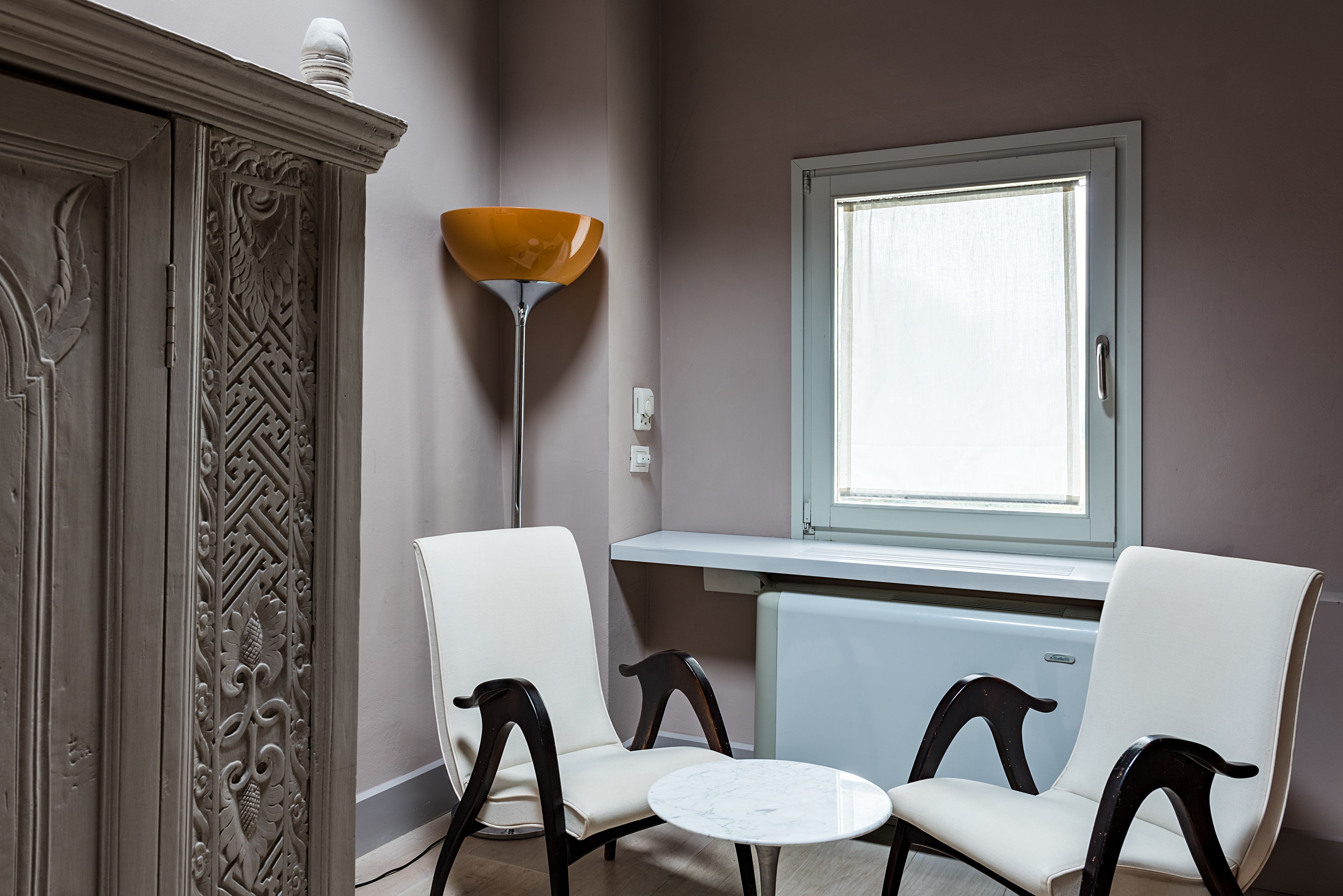 A room with two white chairs, a small round table, a decorative cabinet, a floor lamp, and a window with a view.