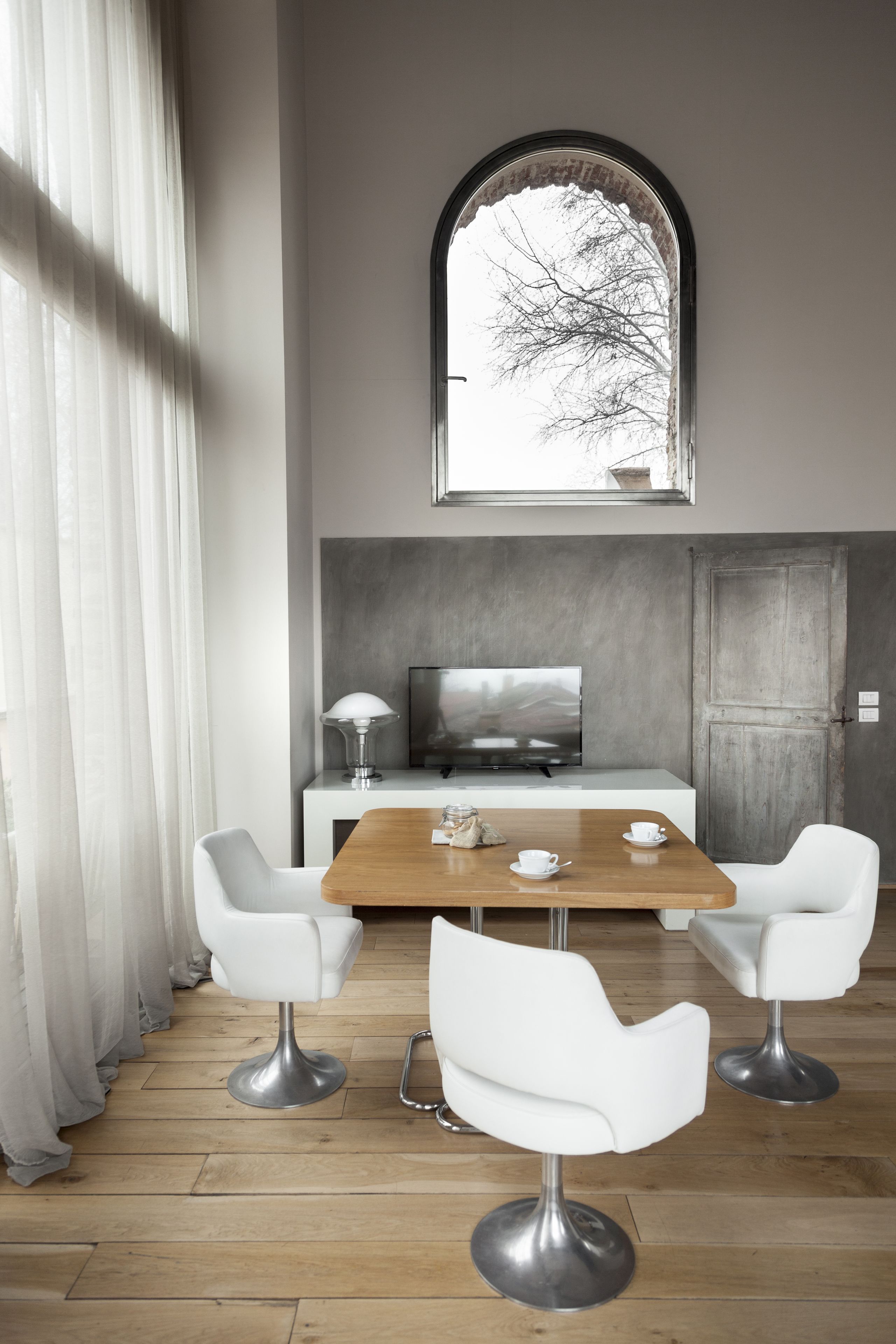 A modern dining area with a wooden table, white chairs, a television, and a window with a view of trees.