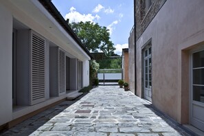 A stone-paved courtyard with a modern building on one side and a tree-lined fence on the other.