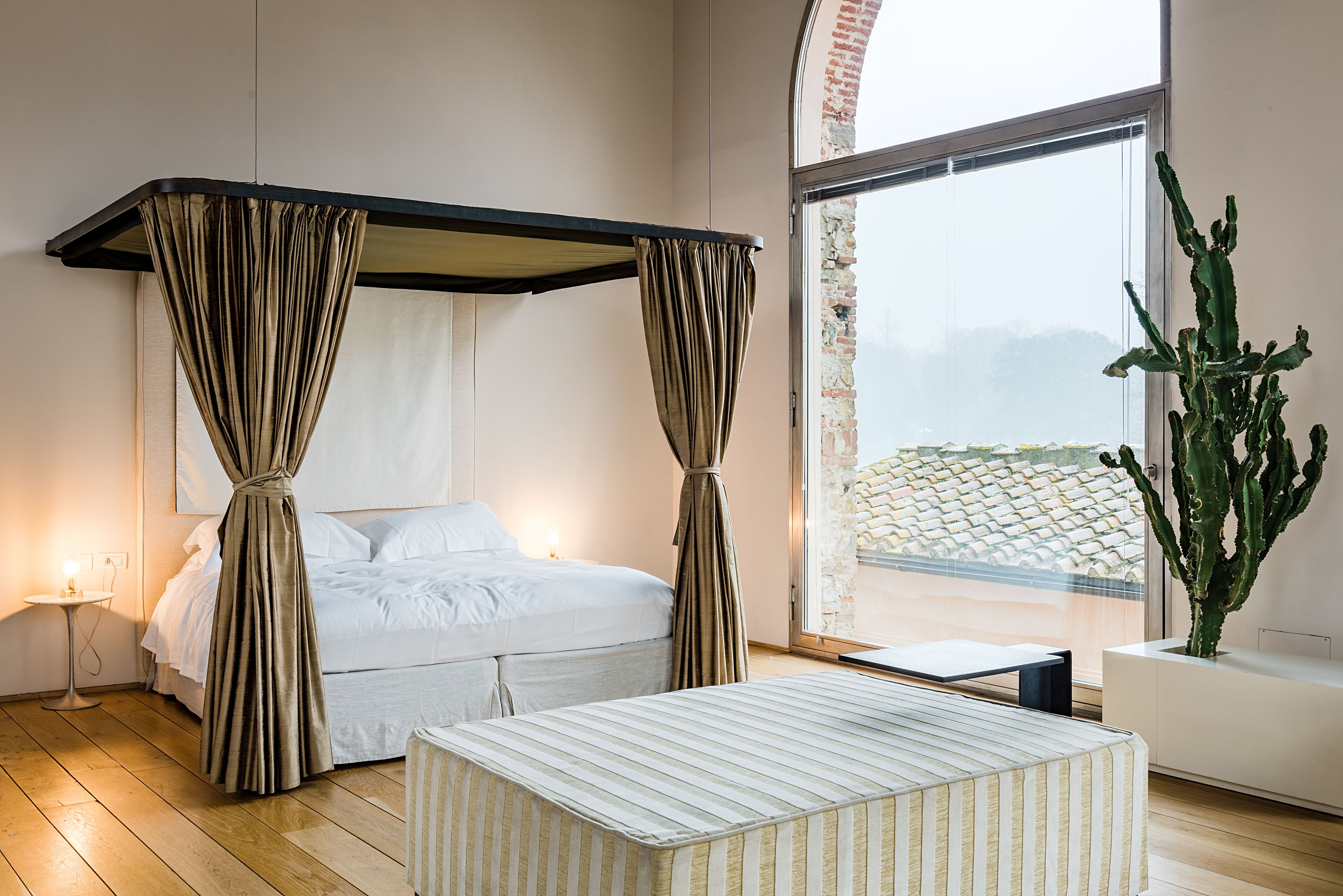 A modern bedroom with a four-poster bed, a bedside table, a potted cactus, and a large window with a view of a stone wall.