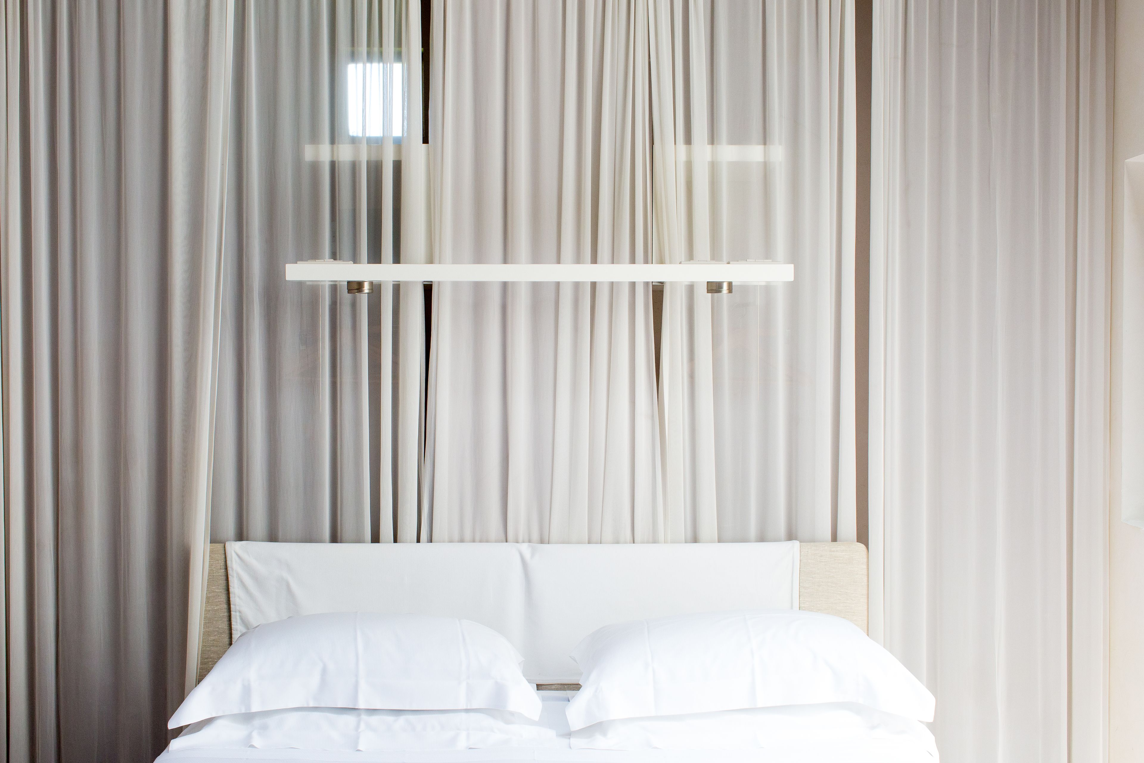 Una cama con sábanas blancas y cabecera de madera, frente a cortinas translúcidas.