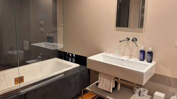 Ein modernes Badezimmer mit einer großen Badewanne, einem Waschbecken und einem Spiegel. Auf dem Waschbecken befinden sich Toilettenartikel.