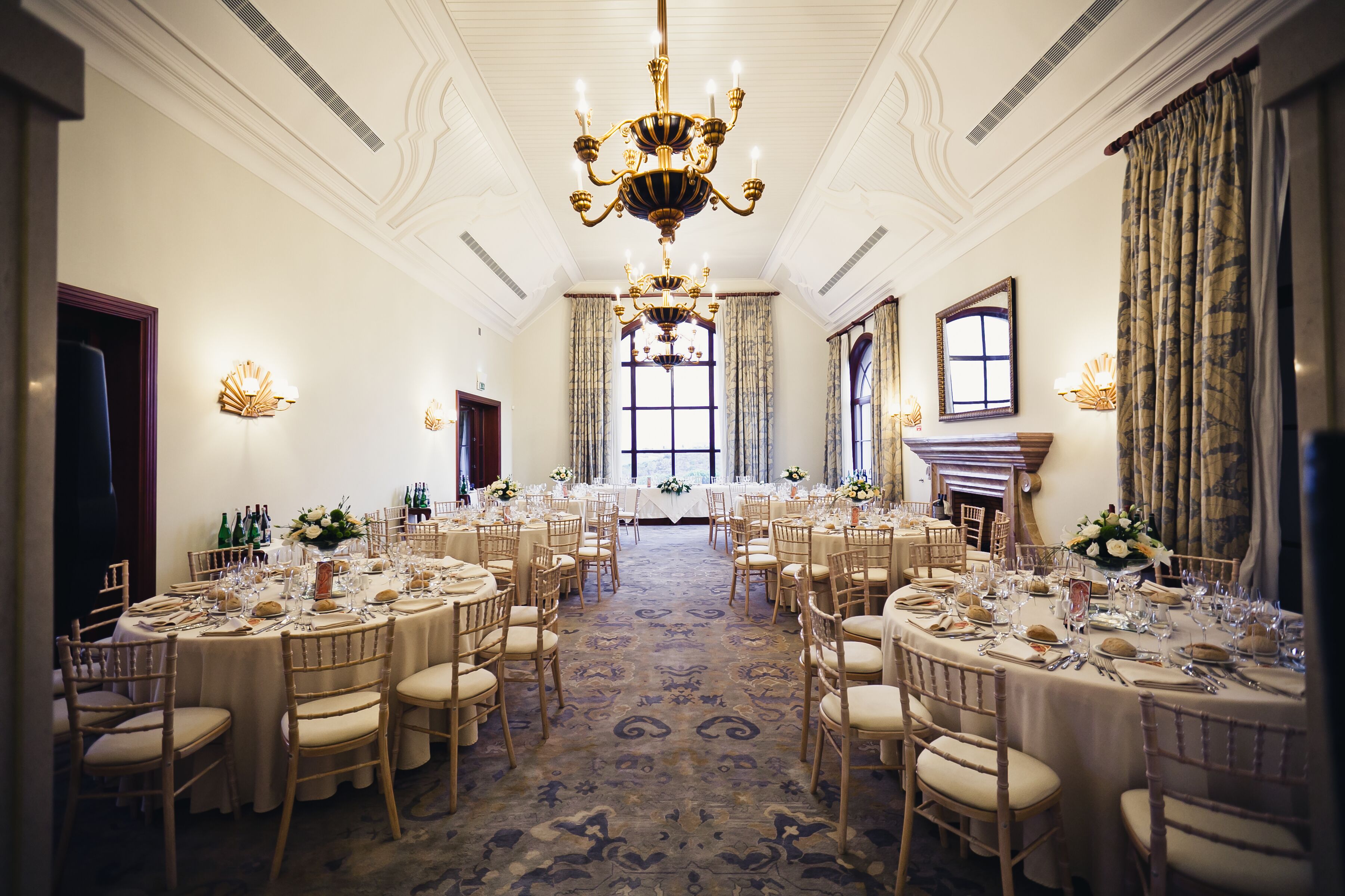A banquet hall with round tables, chairs, chandeliers, and a fireplace.