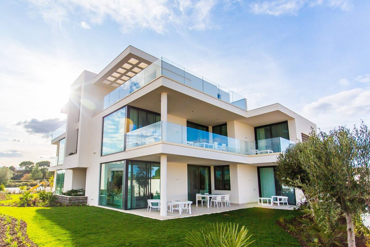 Una casa moderna de dos pisos con techo plano, amplios ventanales y un balcón con mobiliario exterior.