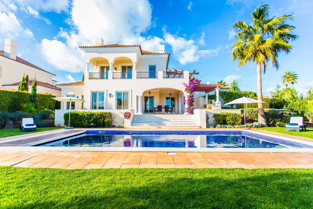 A two-story house with a swimming pool in front, surrounded by well-maintained lawns and palm trees.