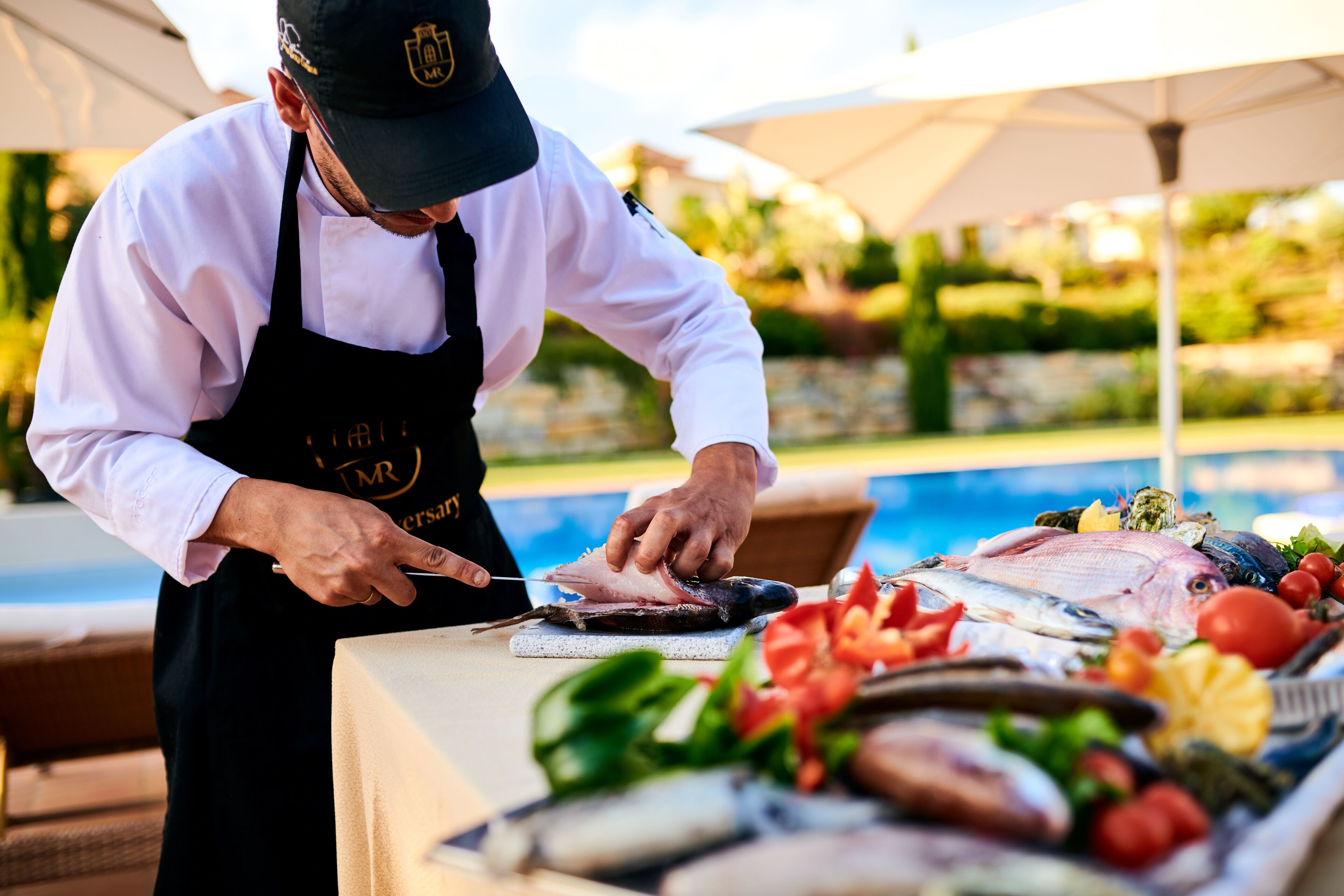 A chef preparing fish at a table with a pool in the background.