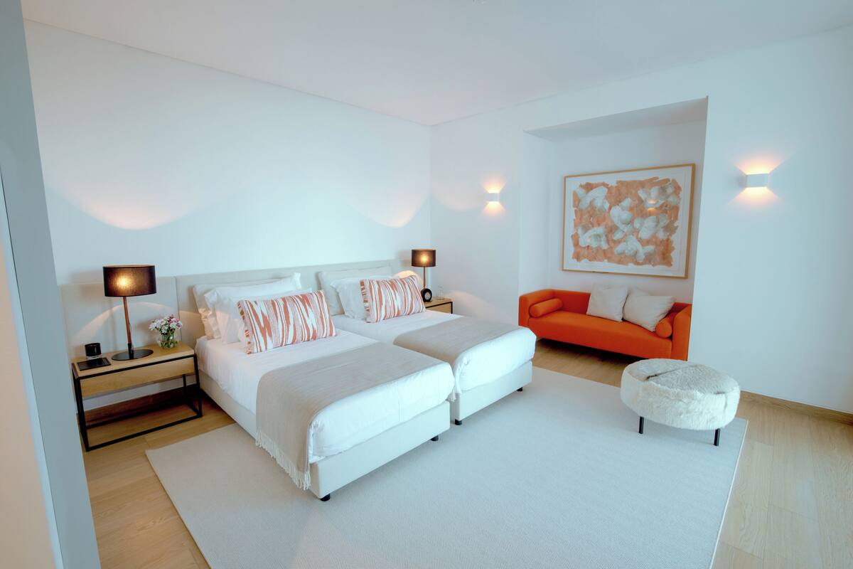 A modern bedroom with a large bed, bedside tables, a nightstand, an orange sofa, and a fluffy ottoman.