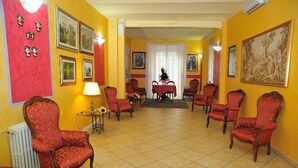 A room with a tiled floor, red upholstered chairs, a table with a vase of flowers, and framed pictures on the walls. - Innocenti (Montecatini Terme)