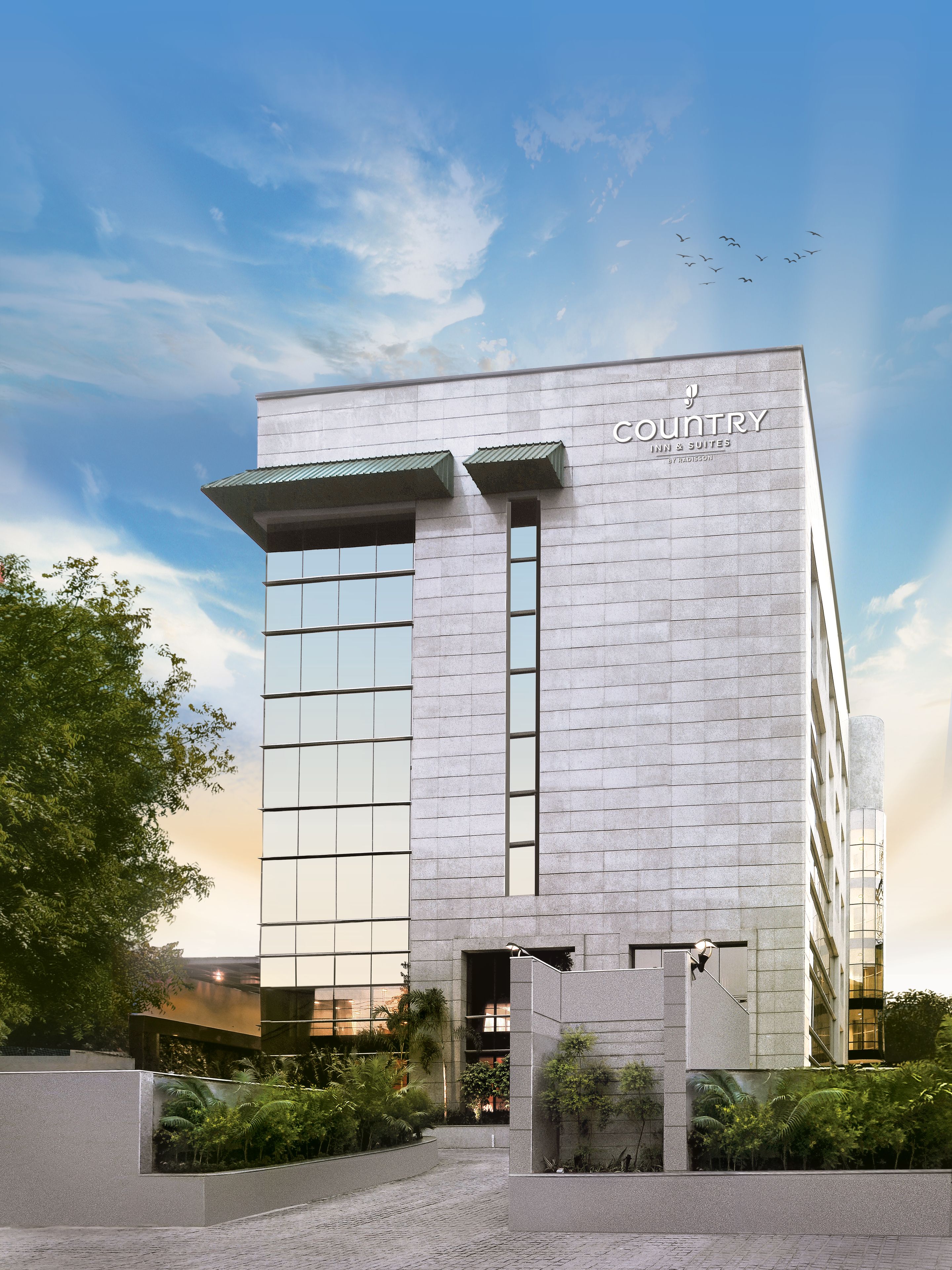 Un edificio moderno con un letrero que dice 'Country Inn & Suites by Radisson'. Cuenta con una entrada destacada con fachada de vidrio y un área ajardinada con follaje.