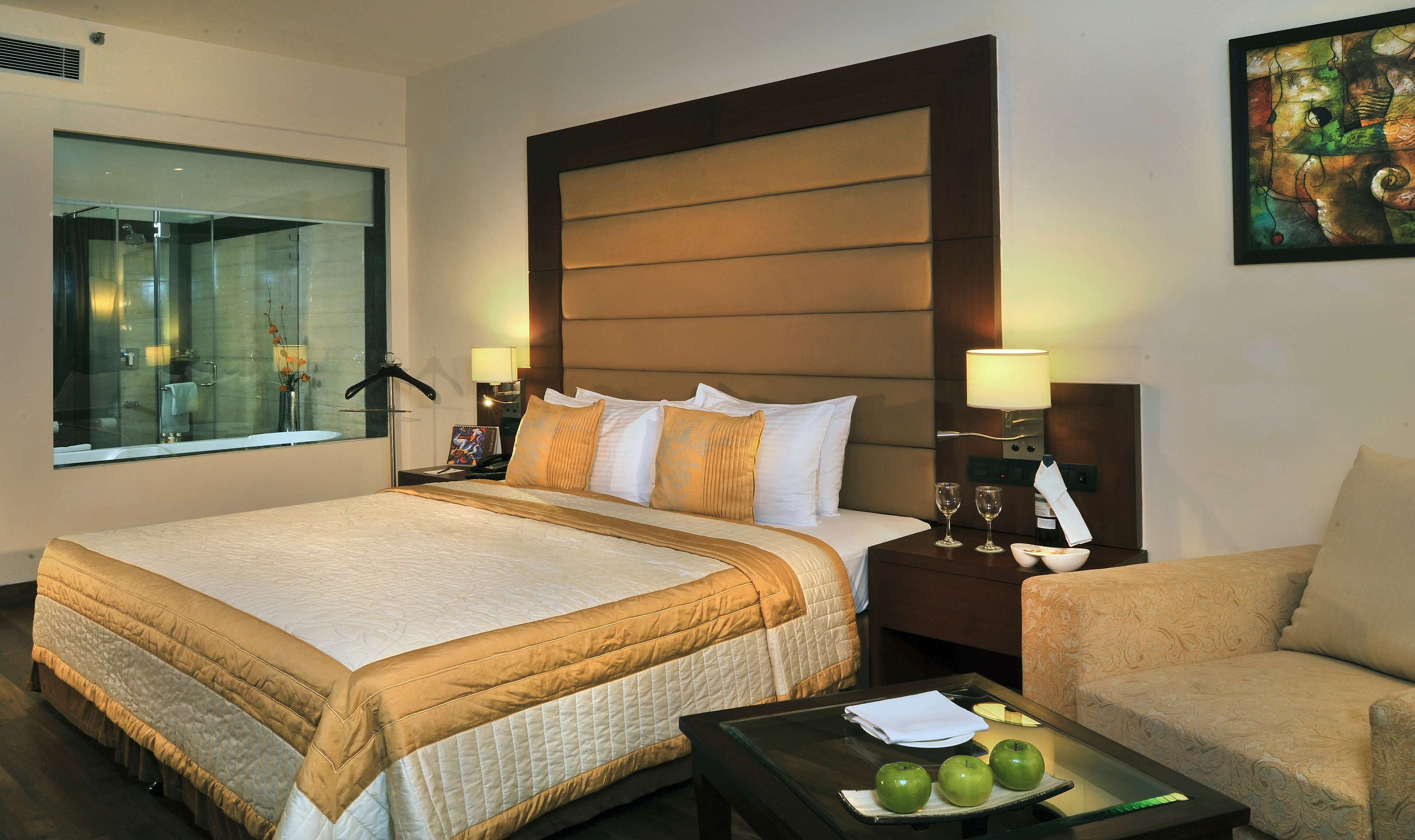 A hotel room with a large bed, a bedside table with a lamp, a chair, and a glass table with apples.