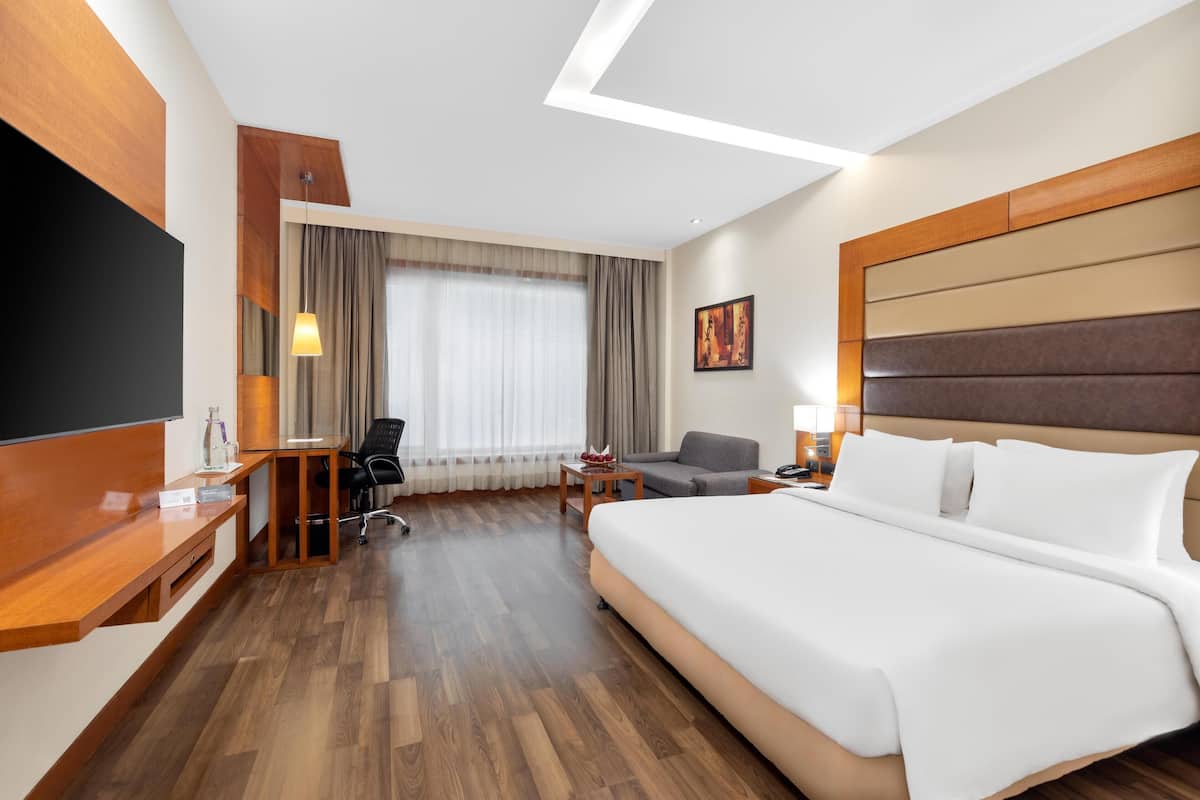 A hotel room with a large bed, a desk with a chair, a sofa, a TV, and a painting on the wall.
