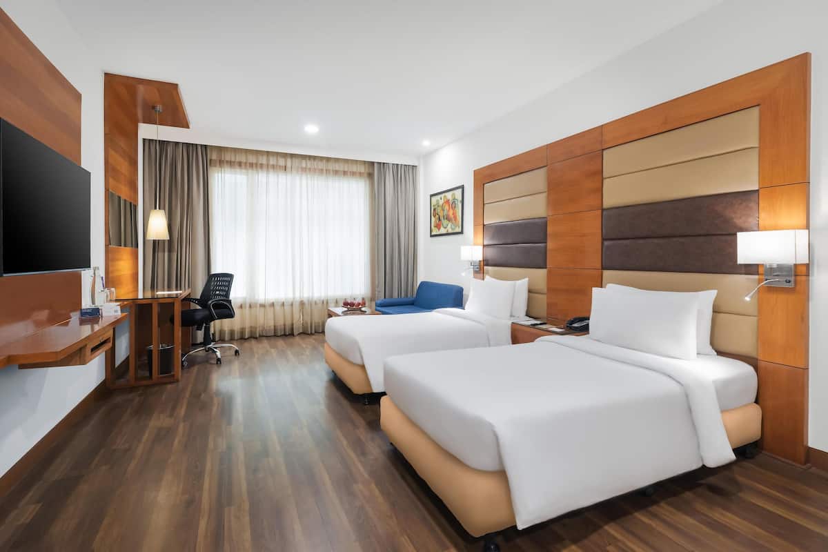 A hotel room with two beds, a desk, a chair, a TV, and a window with curtains.