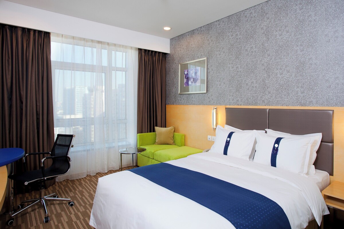 Quarto de hotel com uma cama grande, uma escrivaninha, uma cadeira e vista para a cidade pela janela.