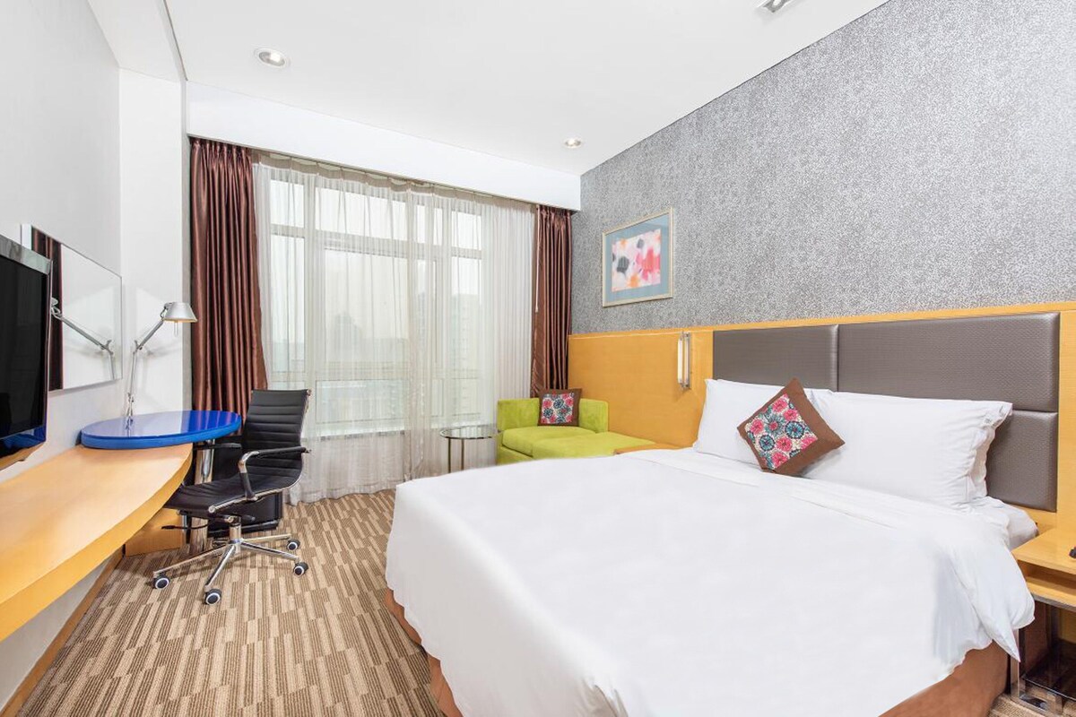 Quarto de hotel com cama, escrivaninha, cadeira, televisão e janela com cortinas.